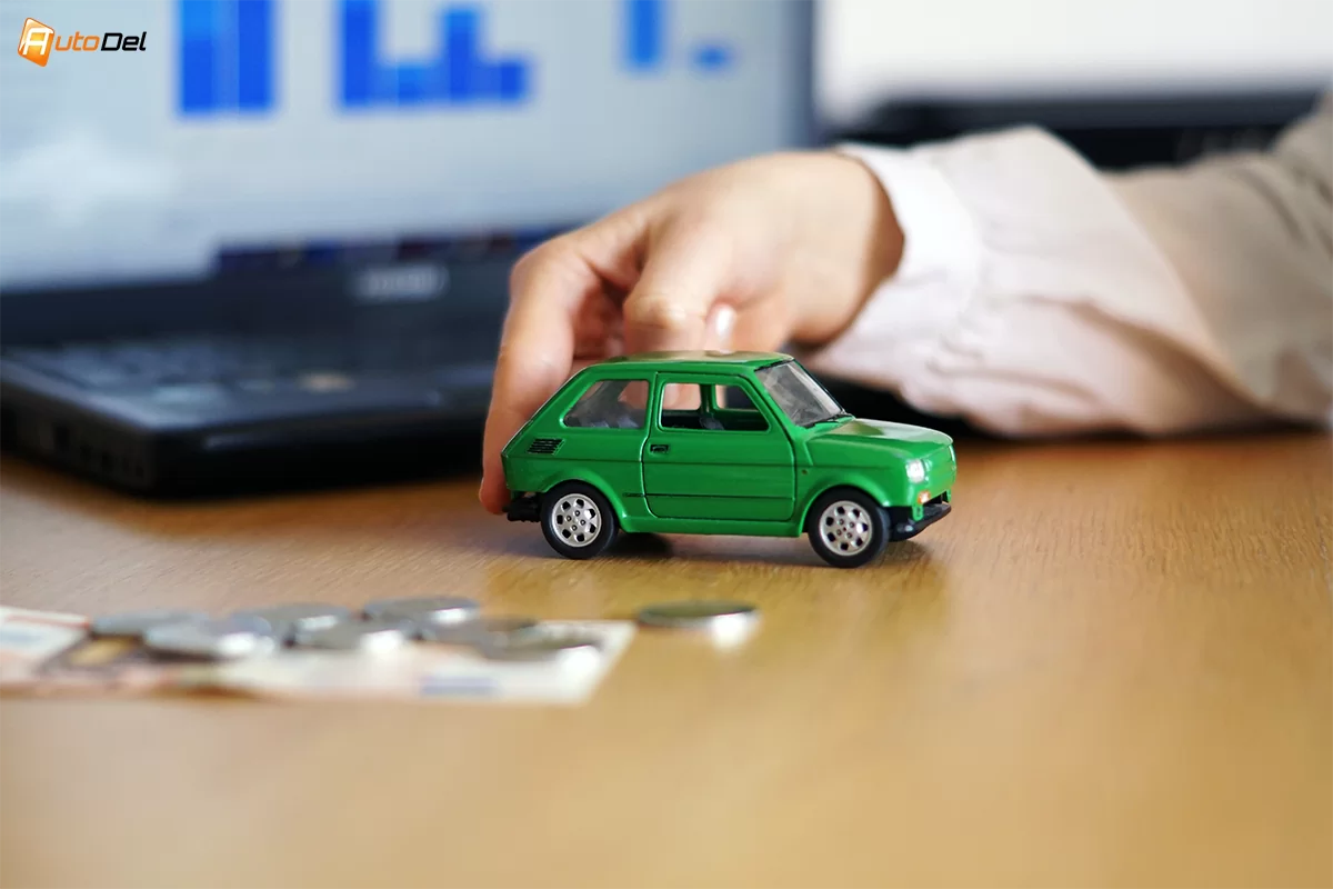 masinuta mica verde impinsa de o mana, cu bani langa si un laptop deschis pe fundal